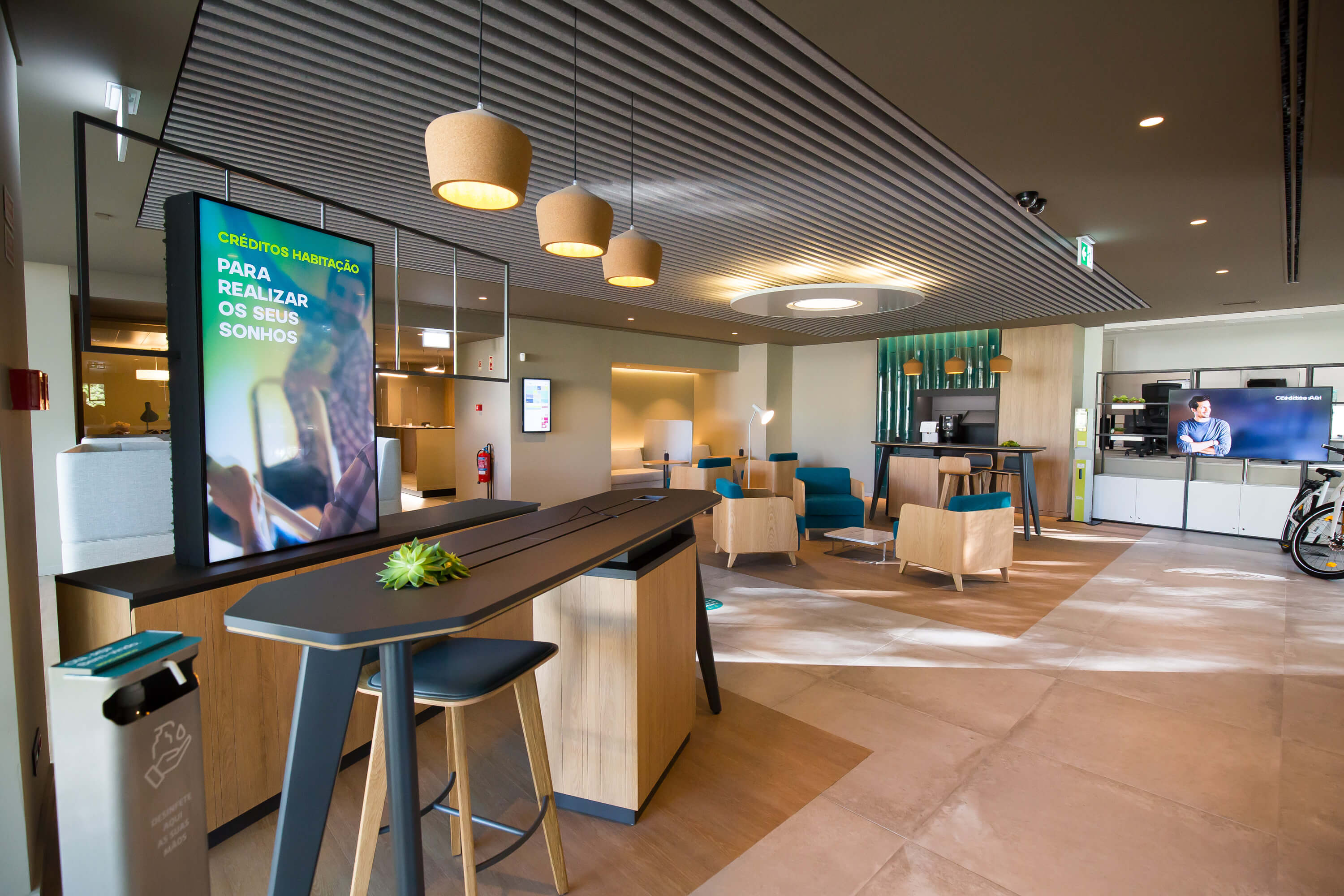 Interior moderno de uma agência novobanco com mesa alta, cadeiras, iluminação suspensa e ecrã digital com mensagem sobre créditos habitação: 'Para realizar os seus sonhos