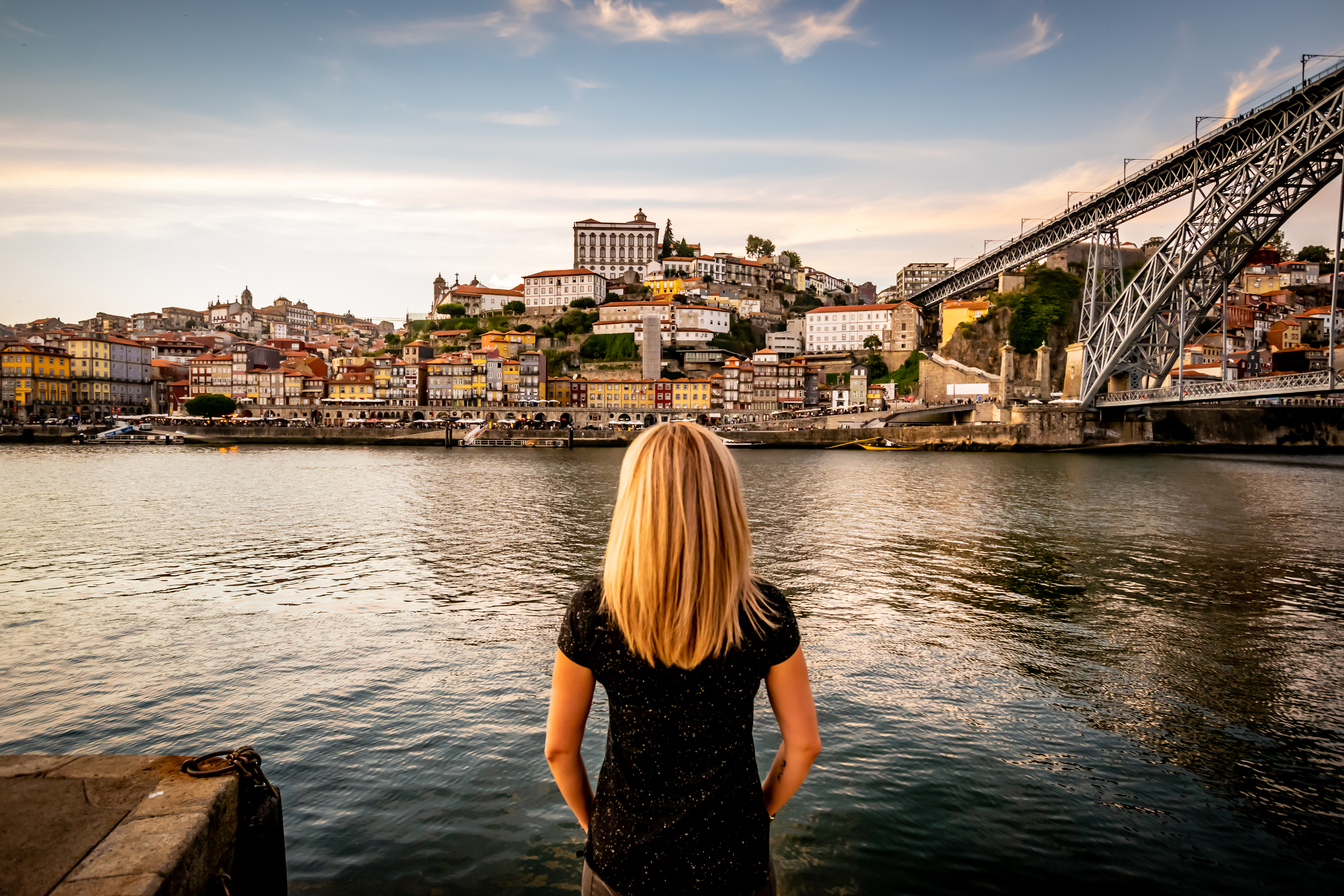 A Portuguese woman admires of the blue sky and soft clouds at sunset displaying Vila Nova de Gaia's colorful City-line from Porto. The double decked metal arched bridge, Don Luís I bridge, overlapping the Douro river, is displayed to the right.